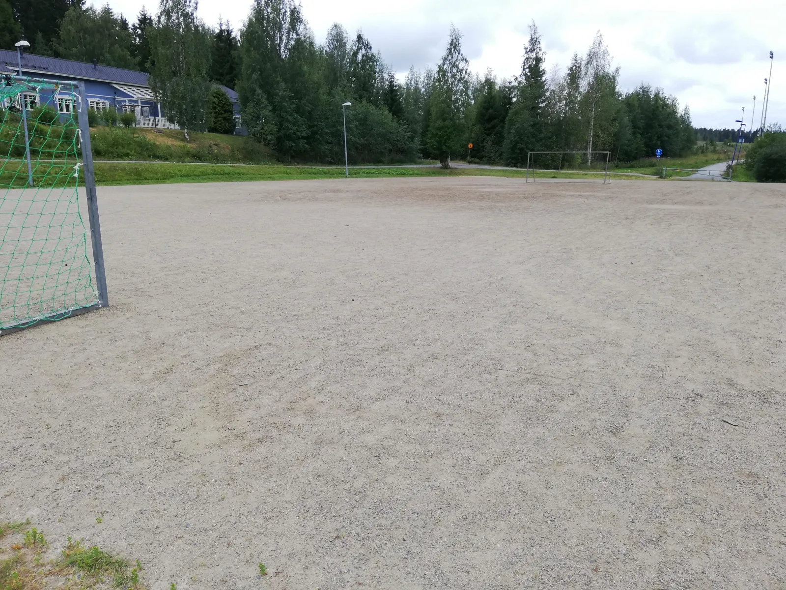 The gravel field beside the Aurinkorinne school in Kuopio, which serves as a free skating field in winter, seen in summer with a goal at the left edge and spruces behind. — Aurinkorinteen luistelukenttä