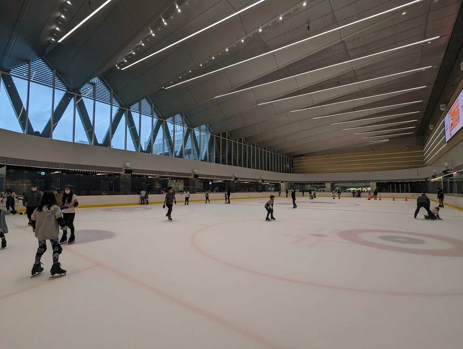 The Aurora Ice Rink sheet under the sloping glass-and-timber roof of the NOKE building, with a few skaters and the centre-ice circles. — Aurora Ice Rink