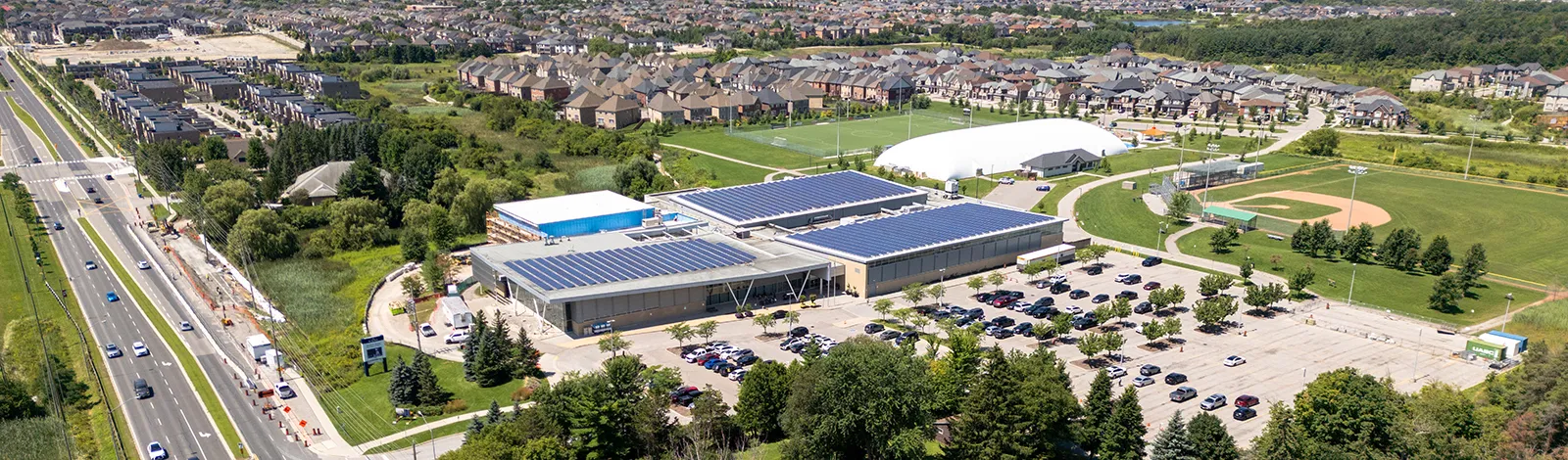 Vista aérea del Aurora Recreation Complex: el largo edificio bajo con paneles solares en el techo, sus estacionamientos y los campos deportivos junto a Wellington Street East. — Aurora Recreation Complex