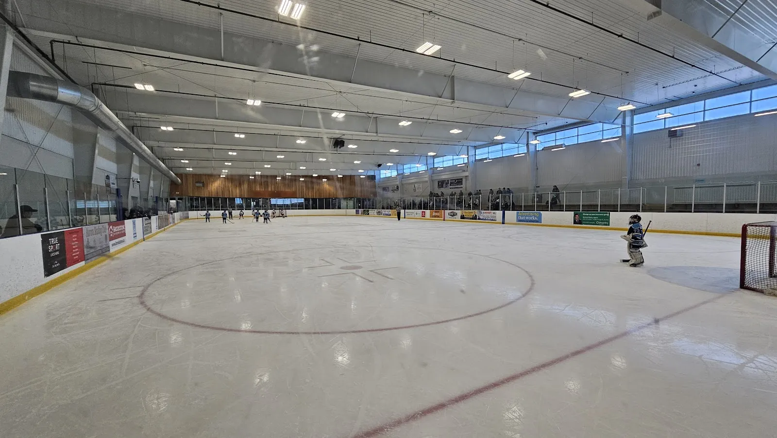 Una pista completa del A.R.C. desde las vallas de un extremo, con un portero en el área cercana, patinadores en el extremo lejano y anuncios de patrocinadores a lo largo de las vallas. — Aurora Recreation Complex