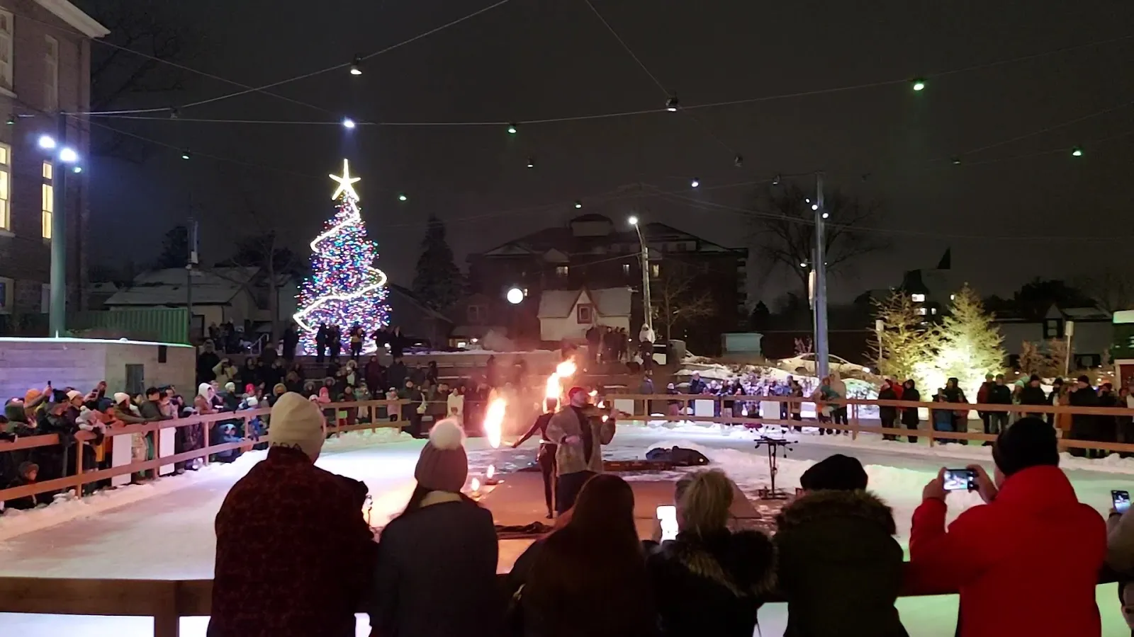An evening event around the Aurora Town Square outdoor rink, with crowds along the boards, a fire performer on the ice and a lit tree behind — Aurora Town Square Outdoor Rink