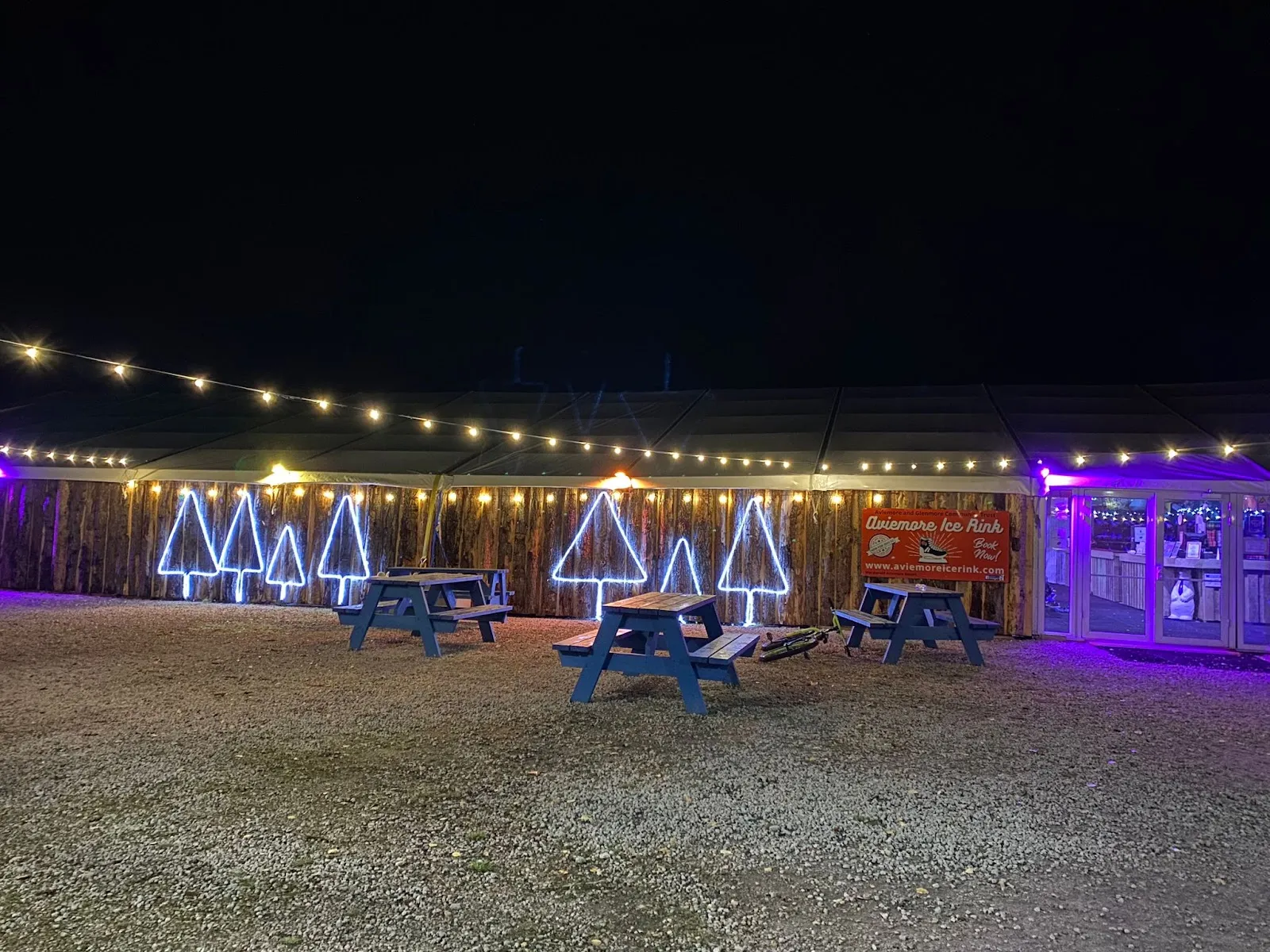 The rink's wood-clad entrance area at night, with the Aviemore Ice Rink sign, strings of lights, light-up fir-tree shapes and picnic benches in front of the marquee. — Aviemore Ice Rink