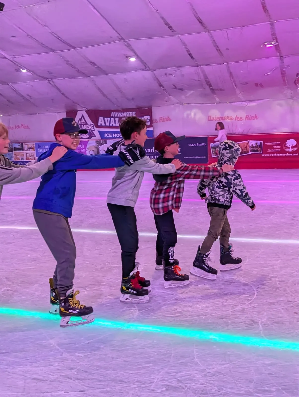Children skating in a chain around the pink-lit tented rink, with the Aviemore Avalanche boards and sponsor panels along the far side. — Aviemore Ice Rink