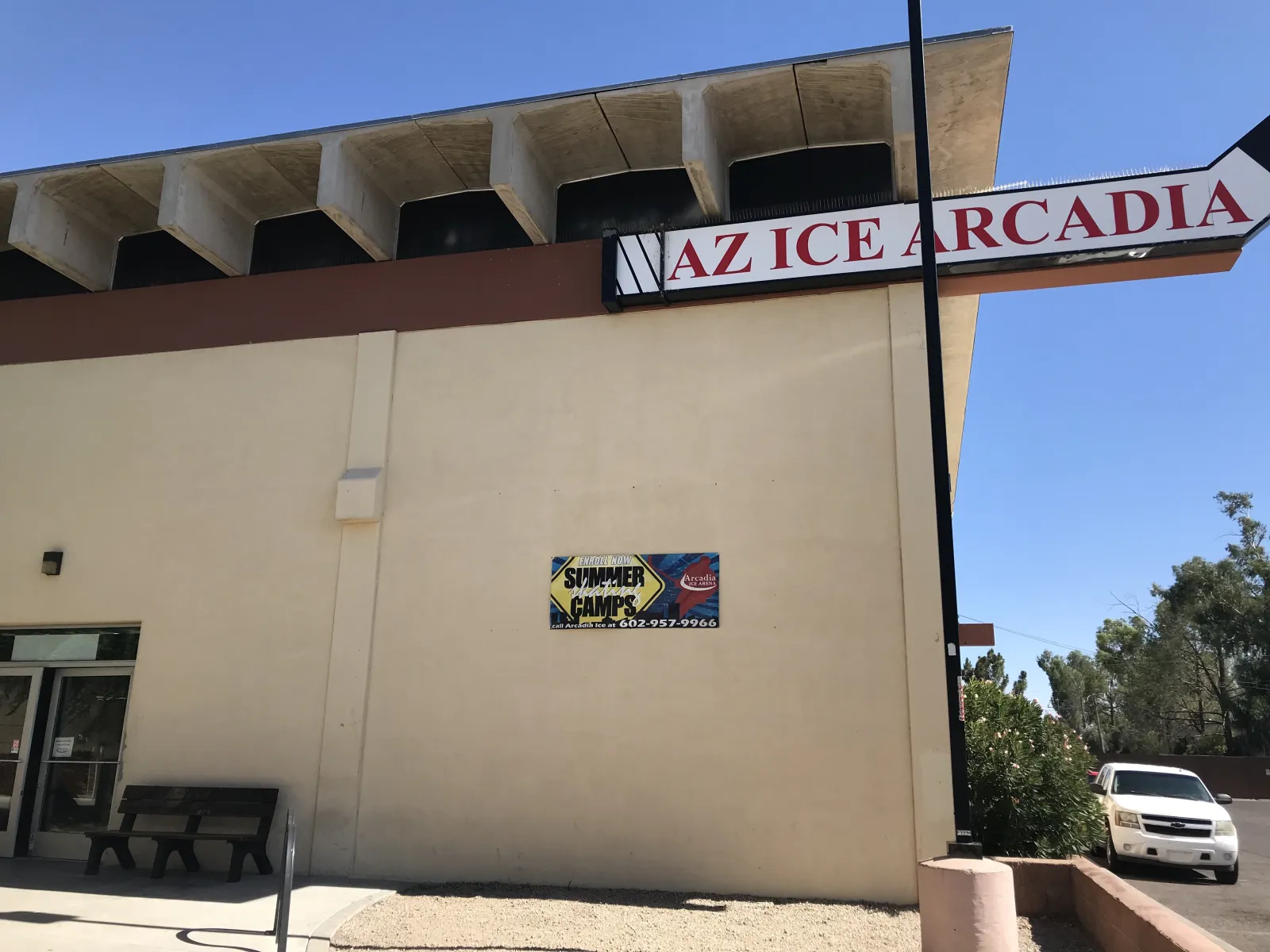 Low angle on the tan stucco corner of the building under a sagging shade canopy, with a red 'AZ ICE ARCADIA' sign mounted on the roofline and a 'Summer Skating Camps' banner on the wall below it; a car and shrubs at street level. — AZ Ice Arcadia