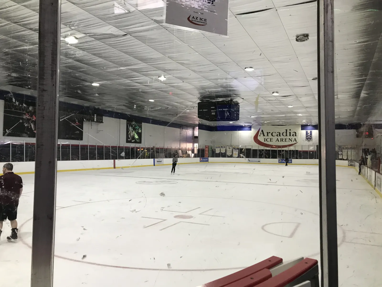 Full ice sheet seen through the glass from the stands, with a red-and-white 'Arcadia Ice Arena' banner and goalie-mask murals on the far wall; one skater near center ice and a couple of people at the boards, otherwise empty. — AZ Ice Arcadia