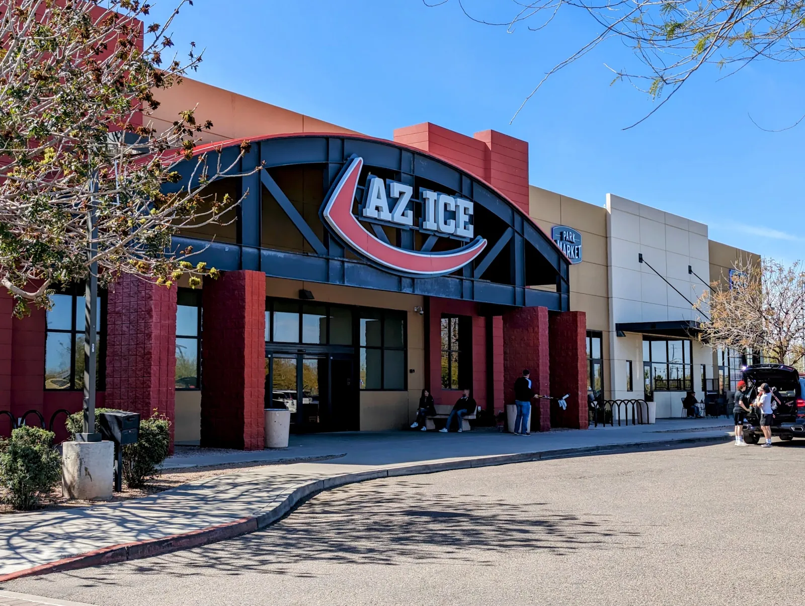 La entrada bajo un gran letrero de AZ Ice en una fachada con arcos rojos y azules, con árboles y jardineras al frente en un día soleado. — AZ Ice Gilbert