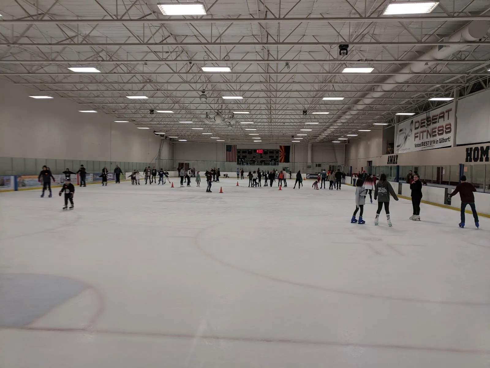 Una sesión pública en una pista amplia, con patinadores repartidos por el hielo bajo un alto techo de acero con un marcador y banderines en la pared del fondo. — AZ Ice Gilbert