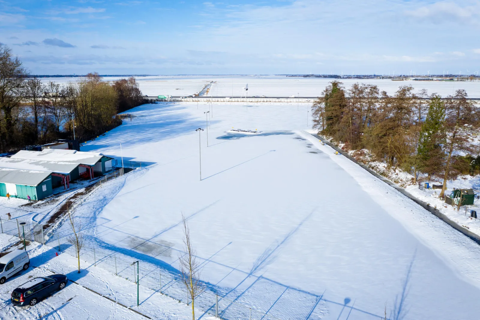 Een luchtfoto van het besneeuwde terrein van de Baarnse baan, met het clubgebouw en een klein parkeerterrein aan de linkerkant en bomen en open velden erachter. – Baarnse IJsbaan