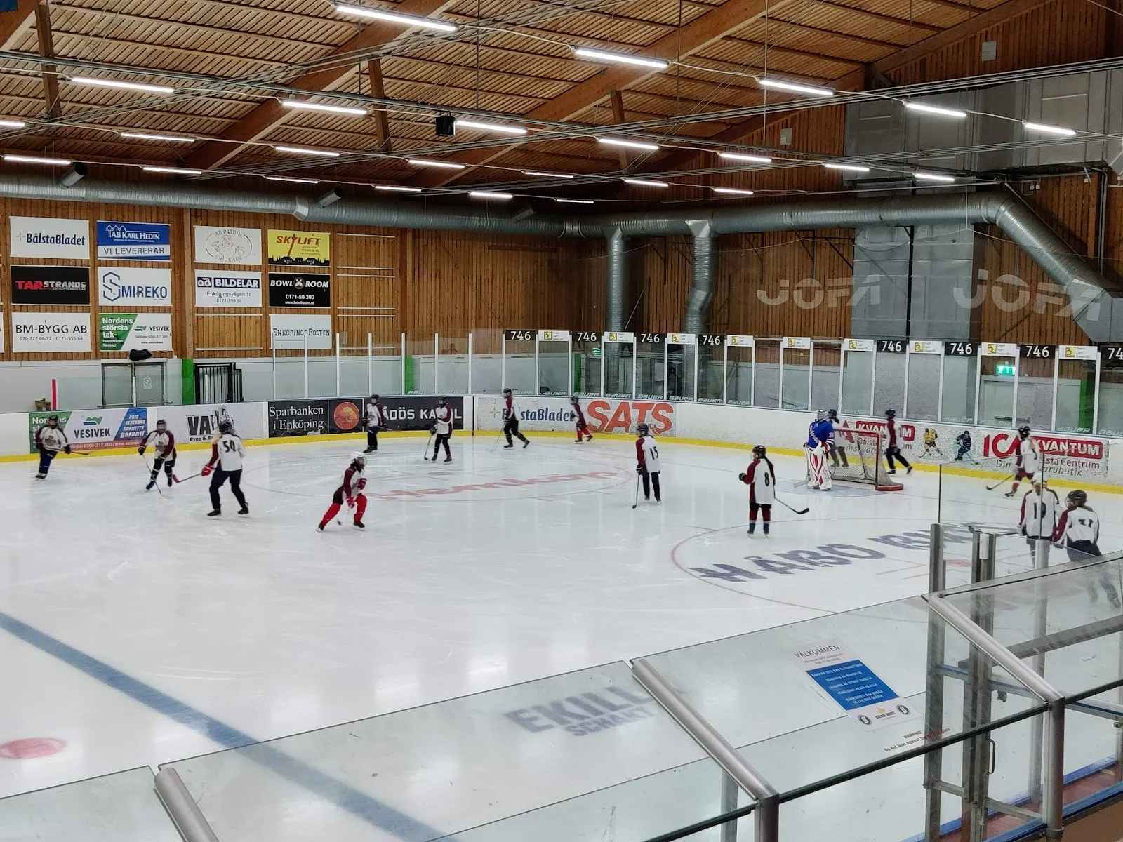 The ice sheet and wooden roof of Bålsta ishall, with a youth practice far off — Bålsta ishall