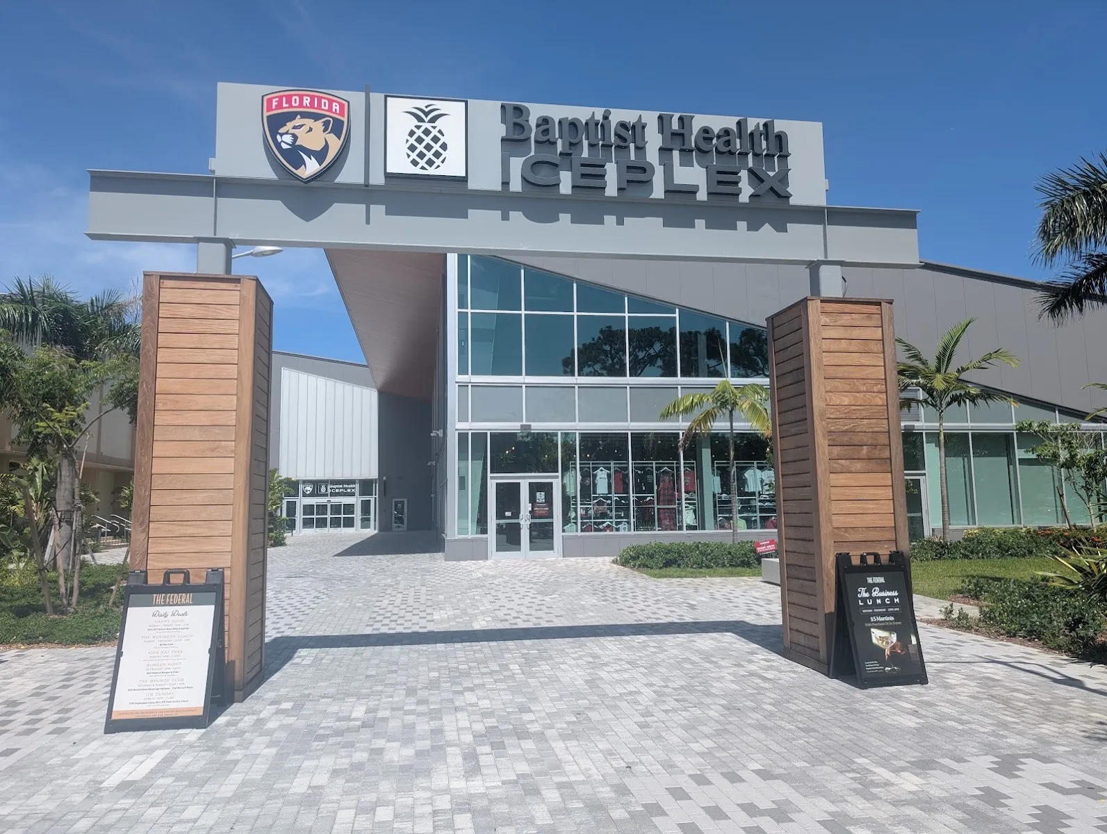 The Baptist Health IcePlex entrance gateway with its Panthers and pineapple signs, wood-clad pillars and a paved plaza in front of the glass doors. — Baptist Health IcePlex (Fort Lauderdale)