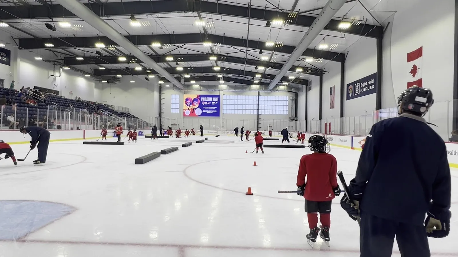 A public session on a wide sheet under a high ceiling, skaters spread across the ice among cones with the IcePlex's tall windows along the far wall. — Baptist Health IcePlex (Fort Lauderdale)