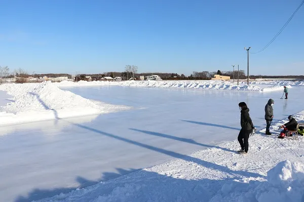 The Baro skating rink in Yubetsu on a clear winter day, a wide sheet of ice banked with plowed snow under a blue sky, with a few people at its edge. — Baro Skate Rink