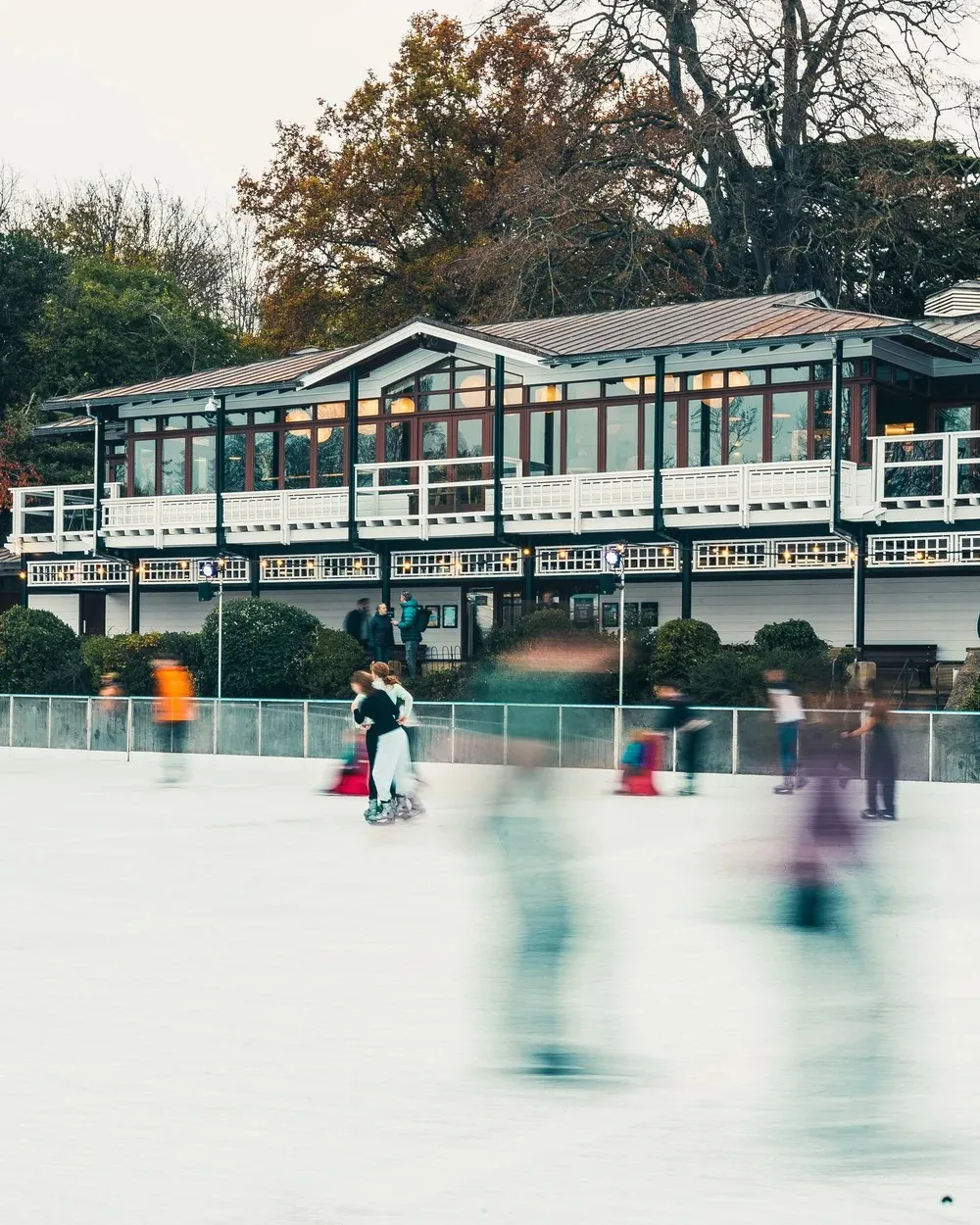 The timber and glass pavilion beside the Bath on Ice rink in Royal Victoria Park, with skaters blurred in front — Bath on Ice