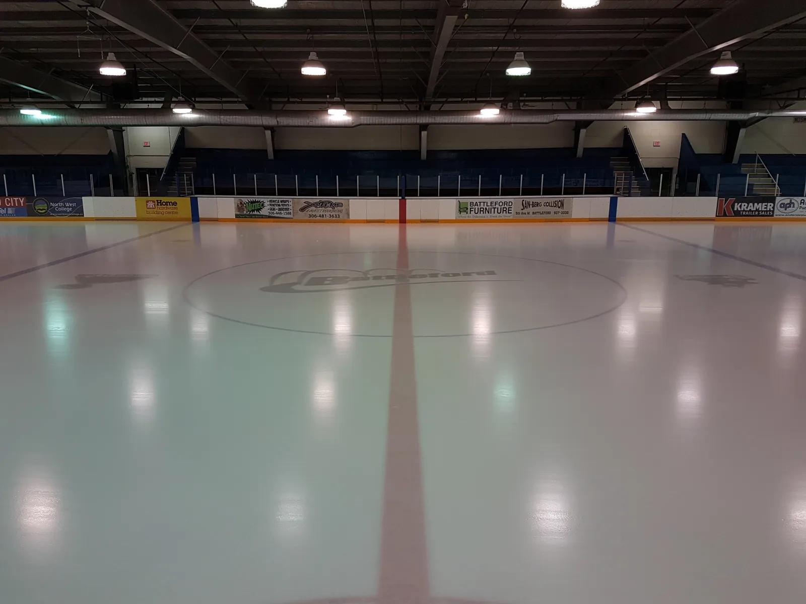 The empty Battleford Arena ice from centre ice, with the Battleford logo in the centre circle and sponsor boards along the far side. — Battleford Arena