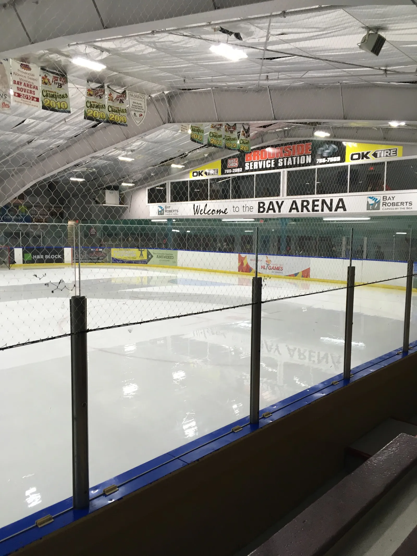 The empty ice sheet of the Bay Arena in Bay Roberts seen over the glass, with the Welcome to the Bay Arena sign above the far boards and green championship banners hanging from the roof. — Bay Arena