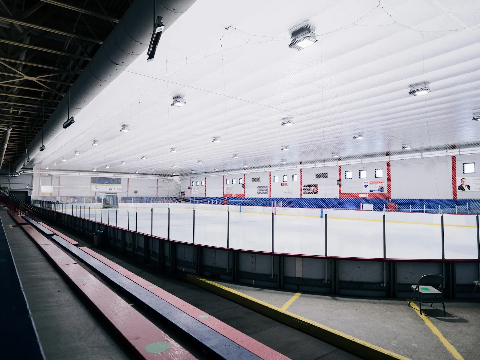 La glace vide vue des gradins, au-delà de la bande, avec des murs rouges et bleus sous un plafond blanc matelassé garni de rangées de lumières. — Beaconsfield Recreation Centre