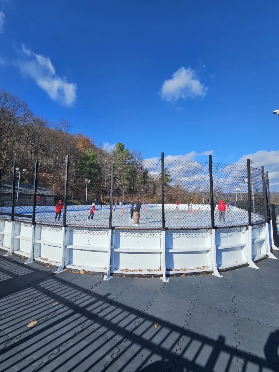 La pista exterior vista desde la terraza junto a las bandas en un día despejado, con patinadores dentro de las bandas blancas y la cerca, y la ladera boscosa detrás. — Bear Mountain Ice Rink