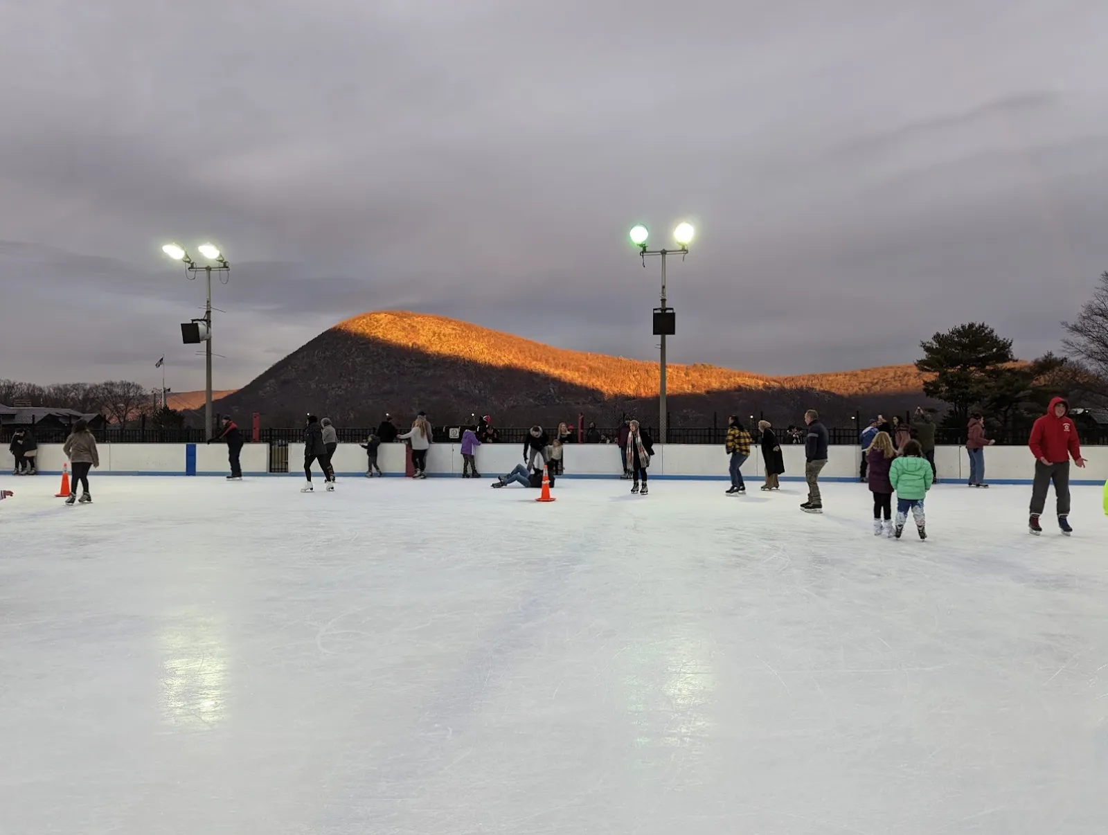 Una sesión pública en la pista exterior al anochecer, con la cima iluminada de Bear Mountain detrás de los patinadores y los reflectores en los postes. — Bear Mountain Ice Rink