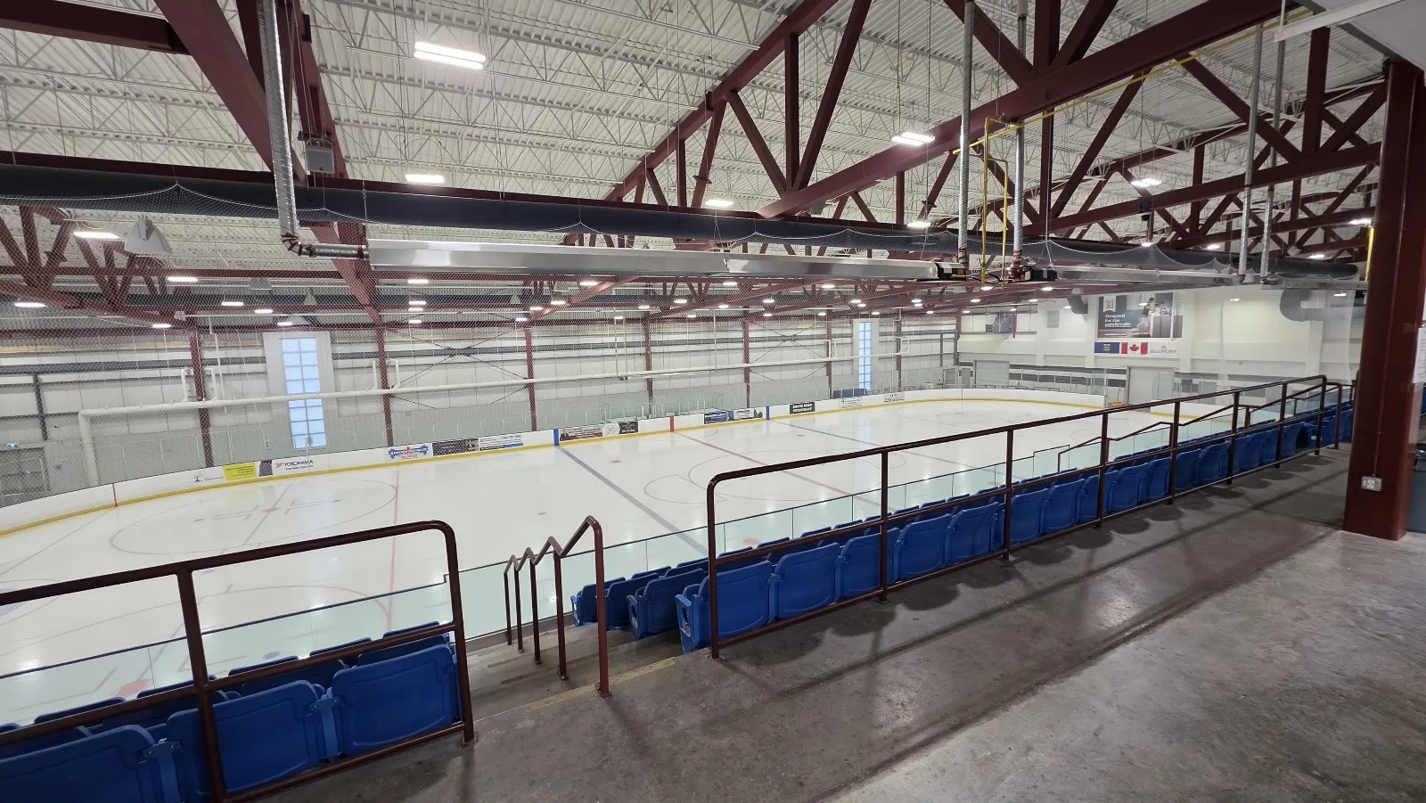 The NHL-size arena from the stands, with blue seats along the boards and the steel roof trusses overhead. — Beaumont Sport & Recreation Centre