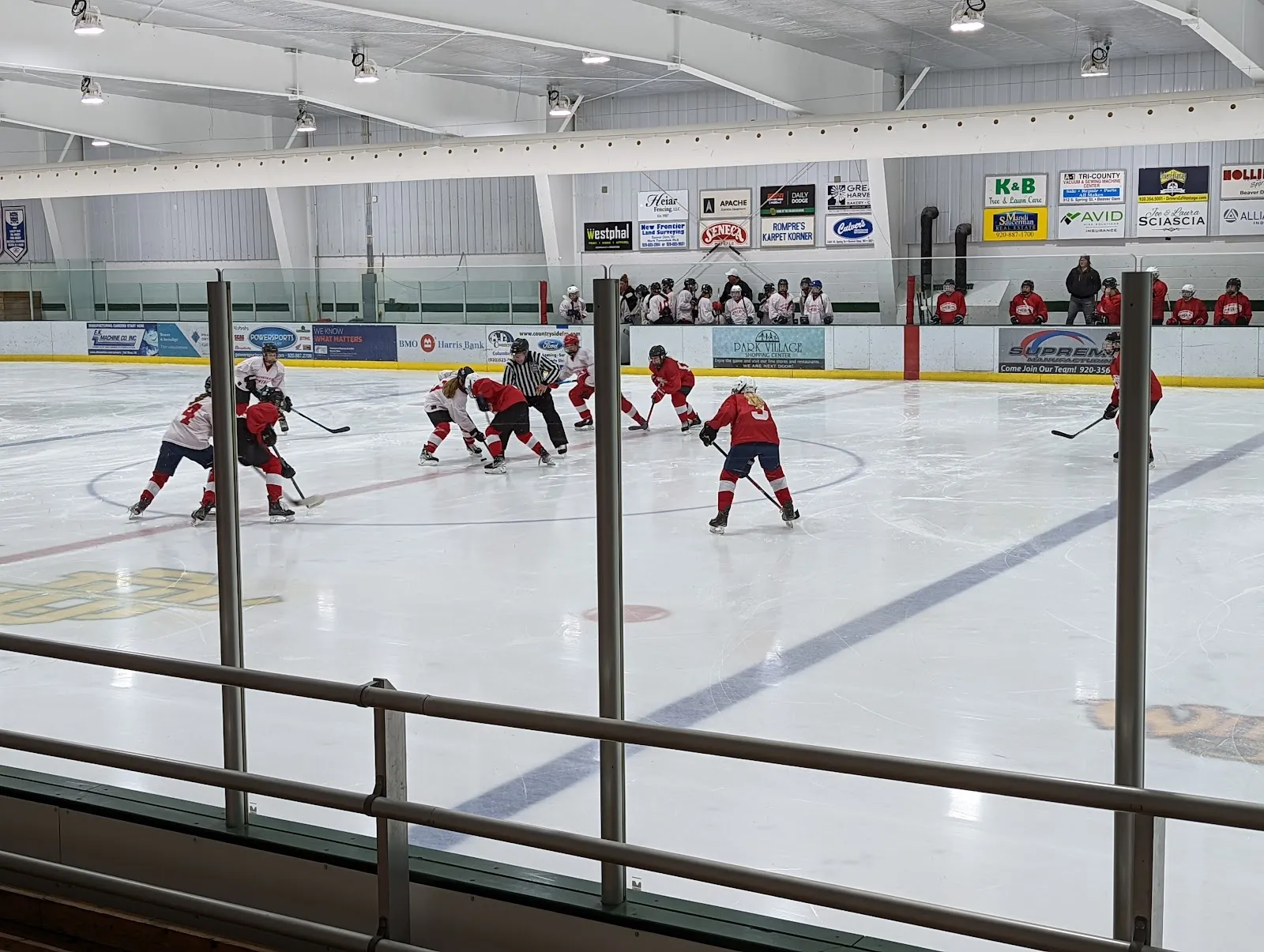 Un partido visto por encima de las vallas, con jugadores repartidos por el hielo y anuncios de comercios locales en la pared del fondo bajo un techo claro. — Beaver Dam Family Center Ice Arena