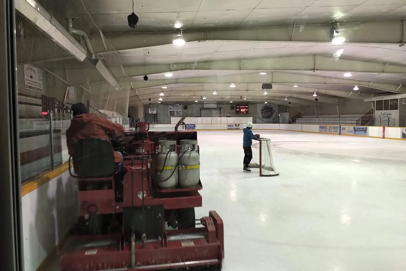 The ice sheet from behind the glass, with the ice resurfacer at work, a skater at the far net and the arena roof and boards. — Beaverlodge Arena