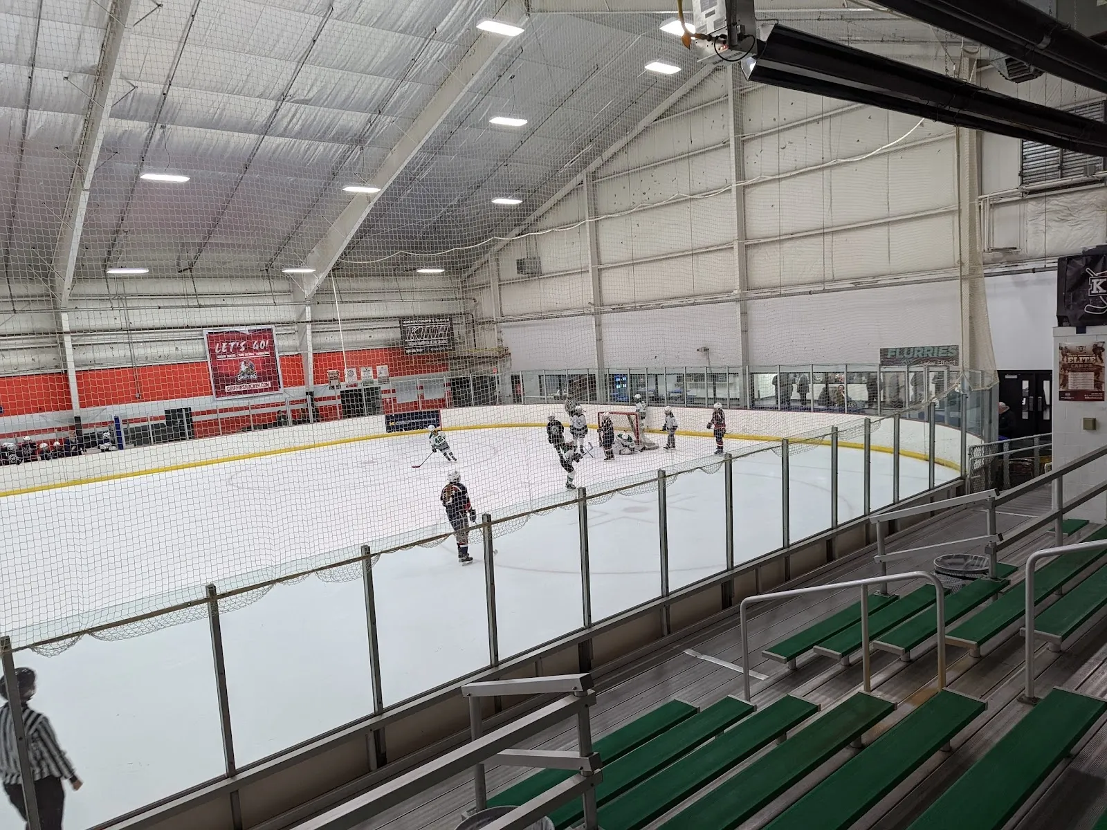 A game seen from the stands beside a blue-railed bench, players and a lone skater spread across the ice under a pale roof with Canadian and Michigan flags on the wall. — Griff's IceHouse Belknap