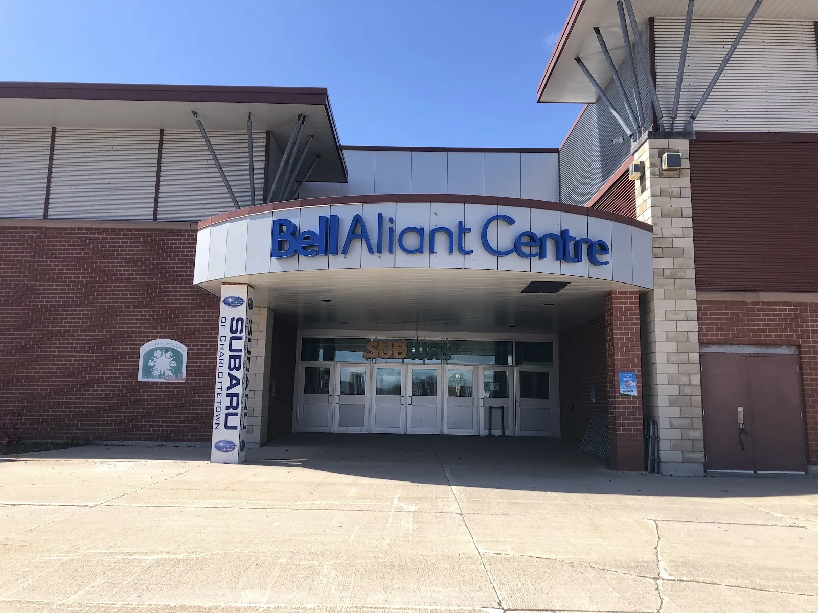 La entrada principal: una marquesina curva con las letras de Bell Aliant Centre sobre puertas de cristal, en un muro de ladrillo rojo bajo un cielo despejado. — Bell Aliant Centre