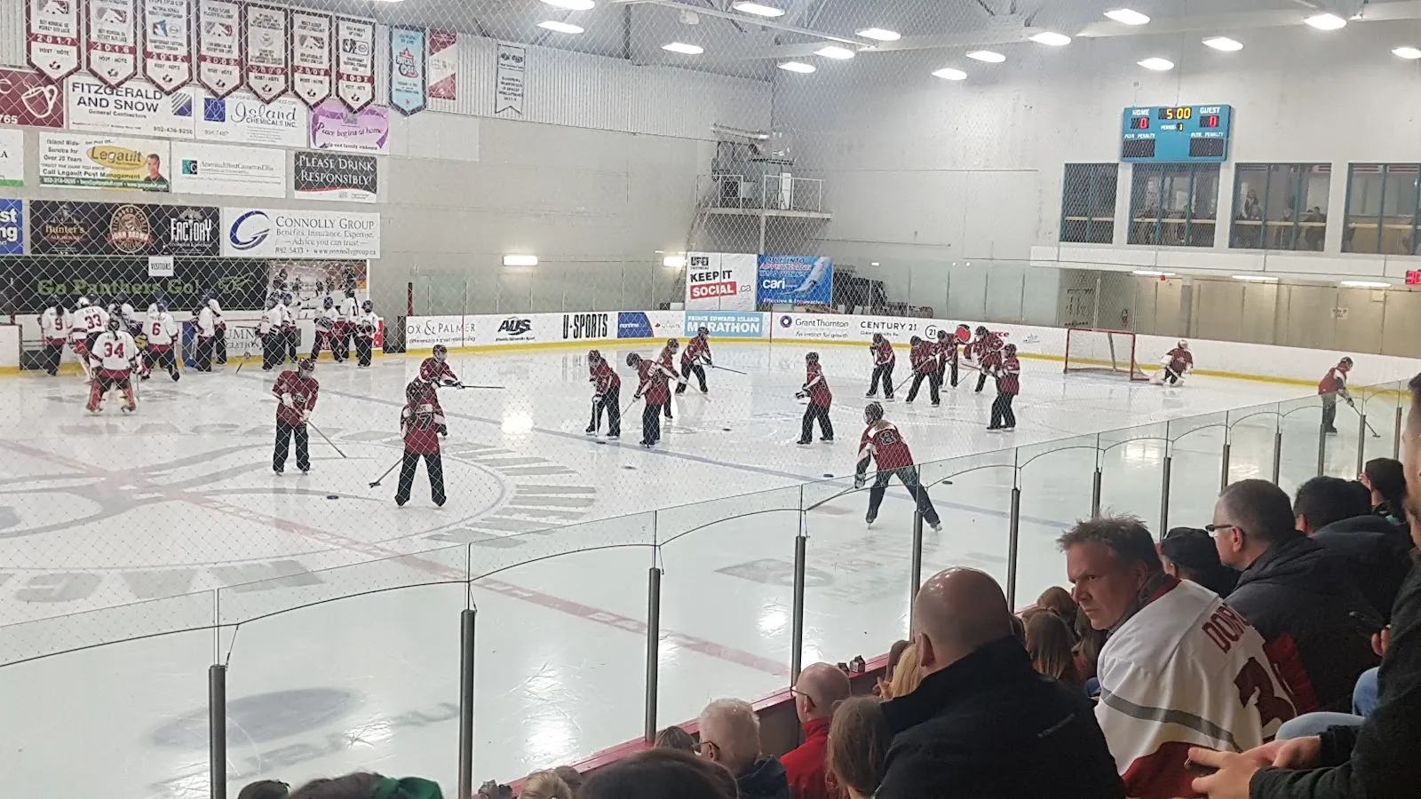 The ice sheet at the Bell Aliant Centre during a game, with players in red and white skating across the faceoff circles, sponsor banners and a scoreboard along the far wall and spectators watching from the stands behind the glass in the foreground. — Bell Aliant Centre