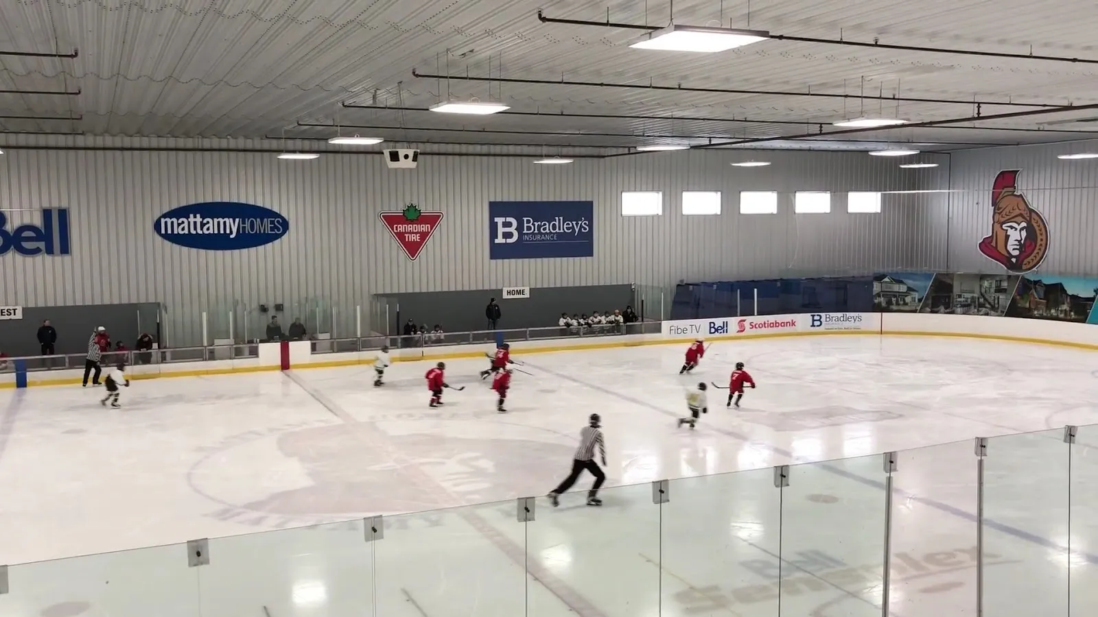 Una pista amplia vista desde las gradas bajas, con jugadores repartidos por el hielo bajo un techo gris con anuncios de Bell y otros patrocinadores y un escudo de los Ottawa Senators en la pared del fondo. — Bell Sensplex