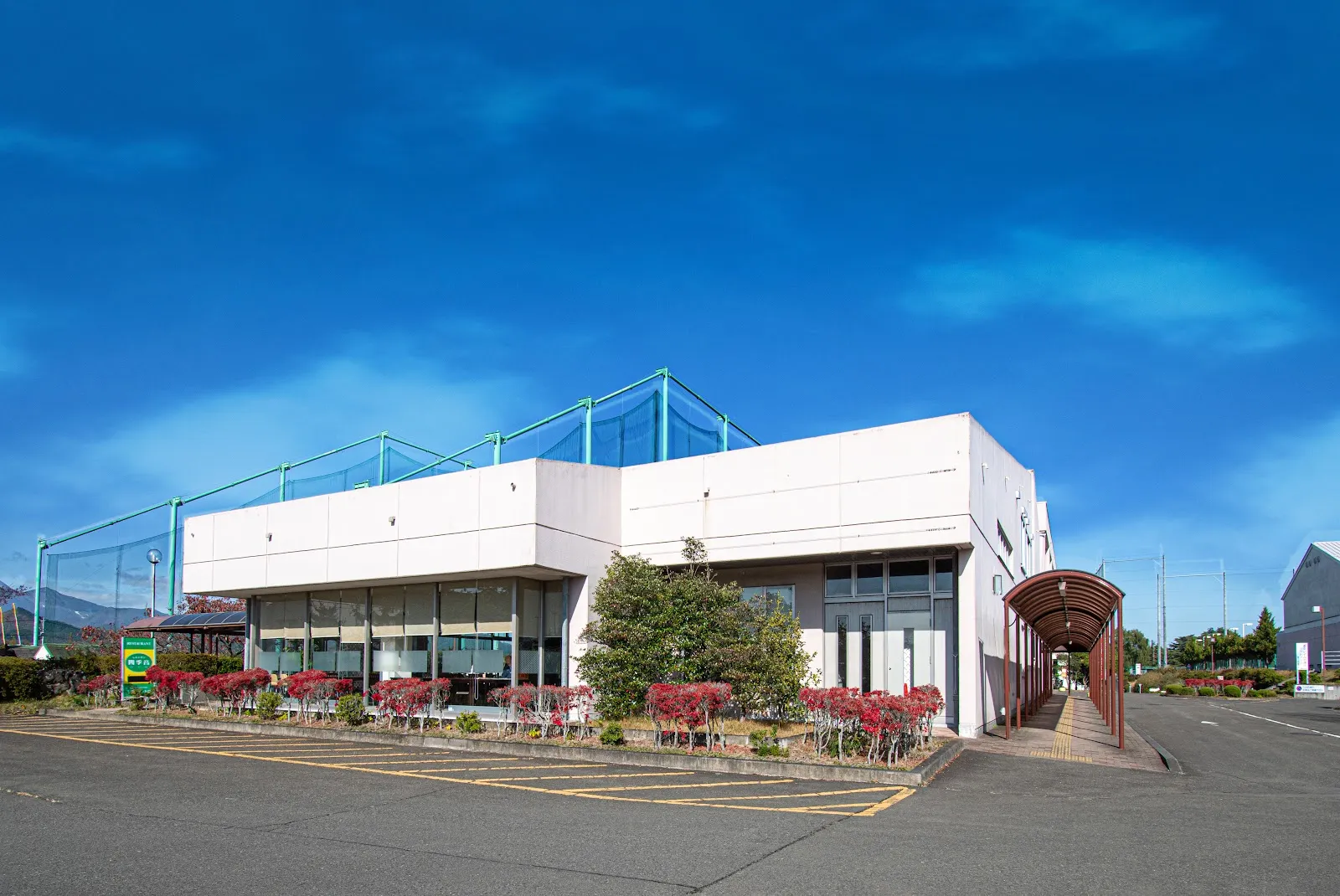 The white two-storey Bell Sunpia building with netting on its roof, red-leaved shrubs along the front and a covered walkway beside the car park under a blue sky. — Bell Sunpia Miyagi Izumi Skating Rink