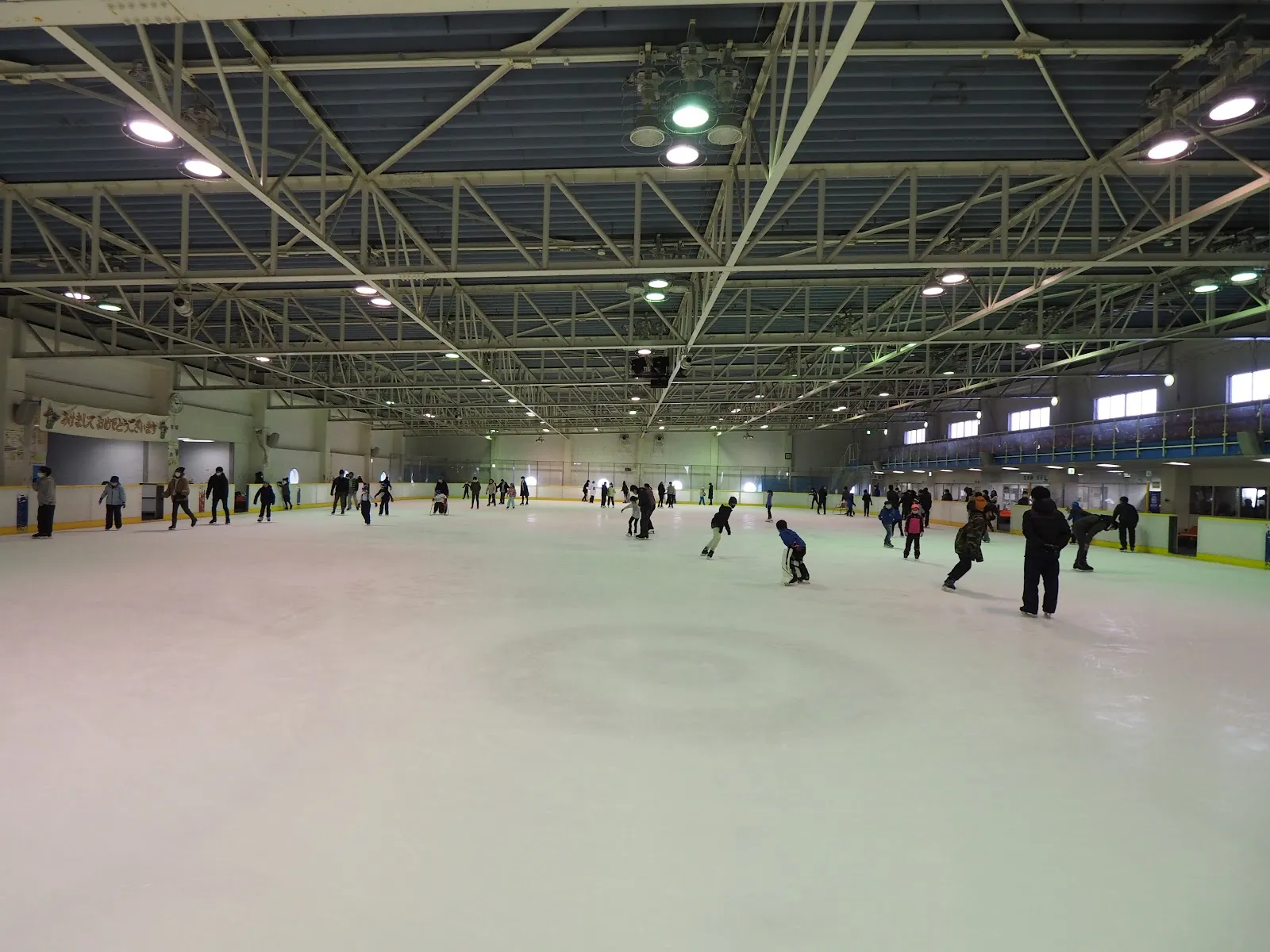 The indoor rink's white ice under a steel-truss roof, with skaters spread across the surface and a banded boards line along the far side. — Bell Sunpia Miyagi Izumi Skating Rink