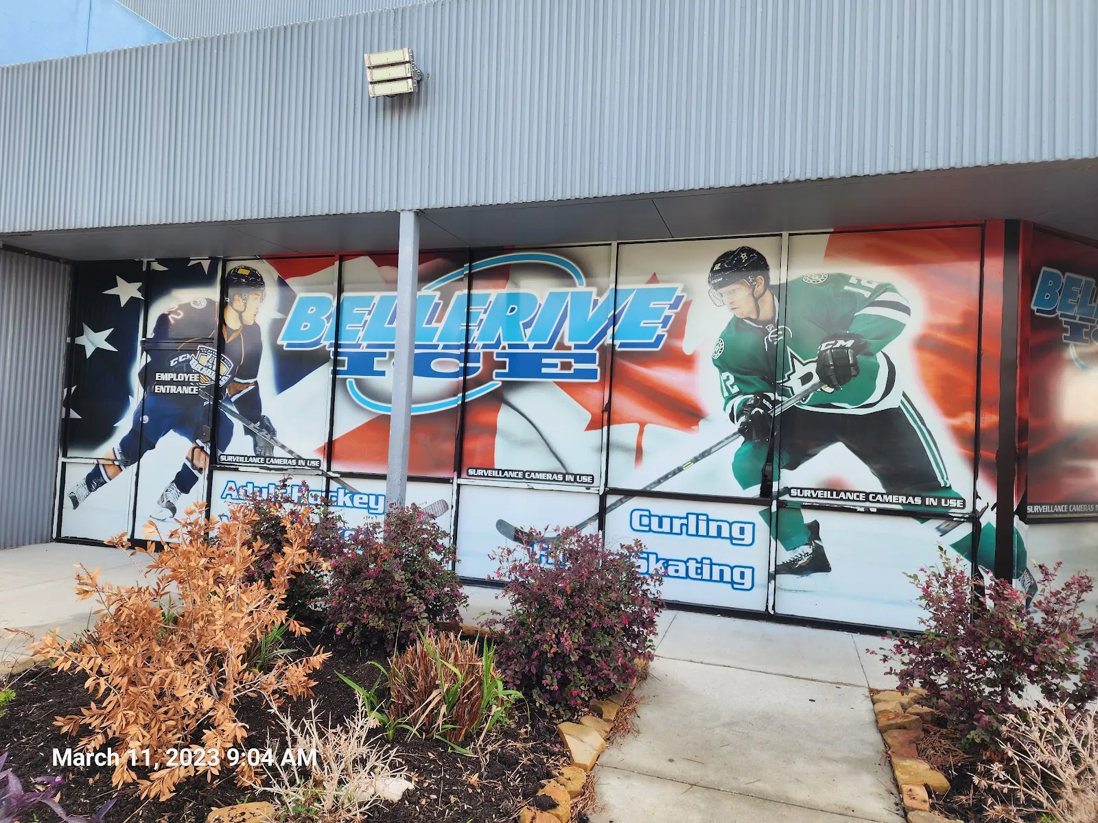 El escaparate de vidrio del edificio hacia la calle, cubierto con grandes gráficos que dicen "Bellerive Ice" flanqueados por ilustraciones de jugadores de hockey y los rótulos "Adult Hockey", "Curling" y "Skating", con arbustos y una banqueta al frente. — Bellerive Ice Center