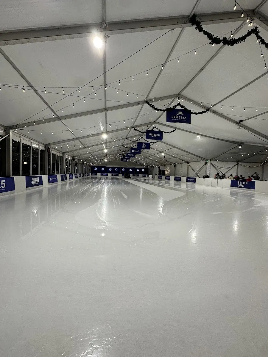 The empty sheet under the white tent roof, strung with lights and hung with sponsor banners, with dark boards along the sides. — Bellevue Downtown Ice Rink