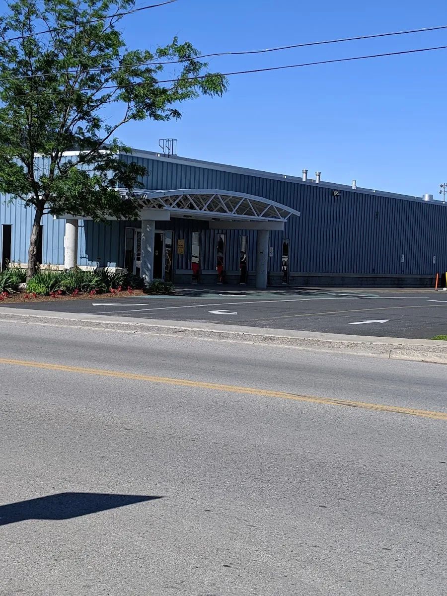La fachada de metal azul grisáceo del Belmont Arena, con una marquesina blanca en arco sobre la entrada, vista desde la calle al otro lado del aparcamiento bajo un cielo despejado. — Belmont Arena & Community Centre (Central Elgin)