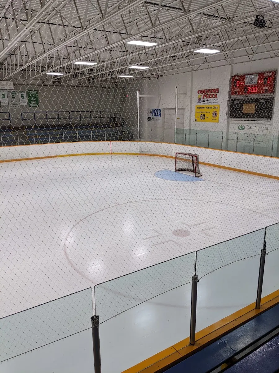The empty Belmont Arena ice under a white steel-truss roof, with the scoreboard and sponsor boards behind the far net. — Belmont Arena & Community Centre (Central Elgin)