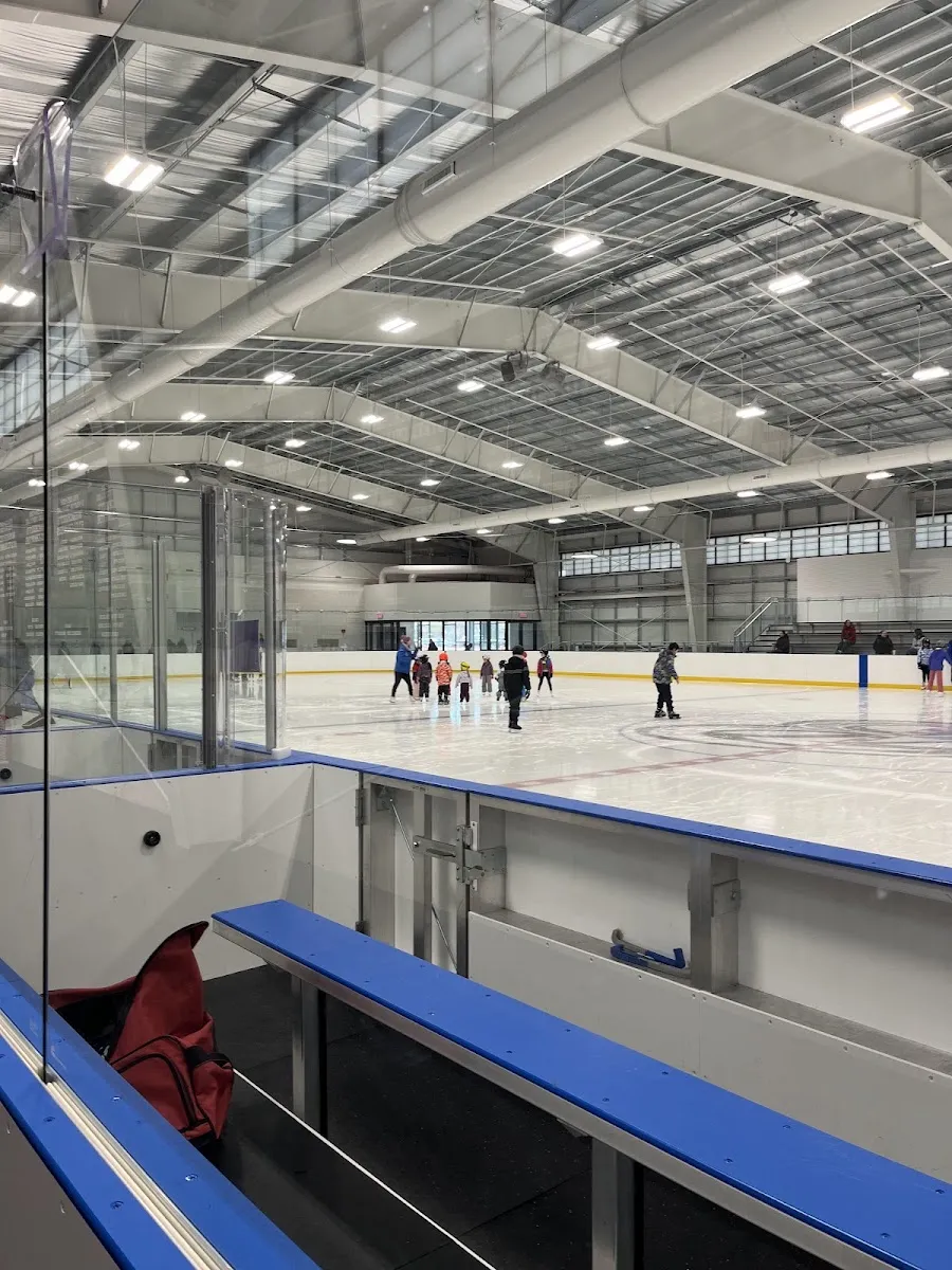 The new rink seen from a player bench: blue-capped boards, glass and a white steel roof, with a few skaters far across the ice. — Belmont Sports Complex