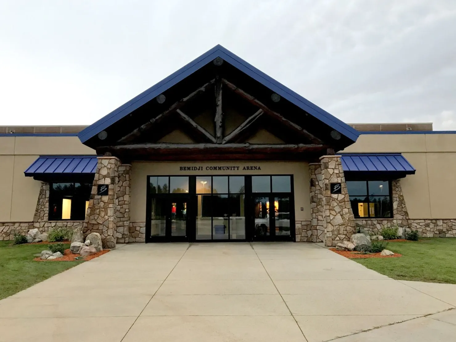 The main entrance, a log-trussed gable with 'Bemidji Community Arena' over the glass doors, fieldstone pillars and blue awnings on either side. — Bemidji Community Arena