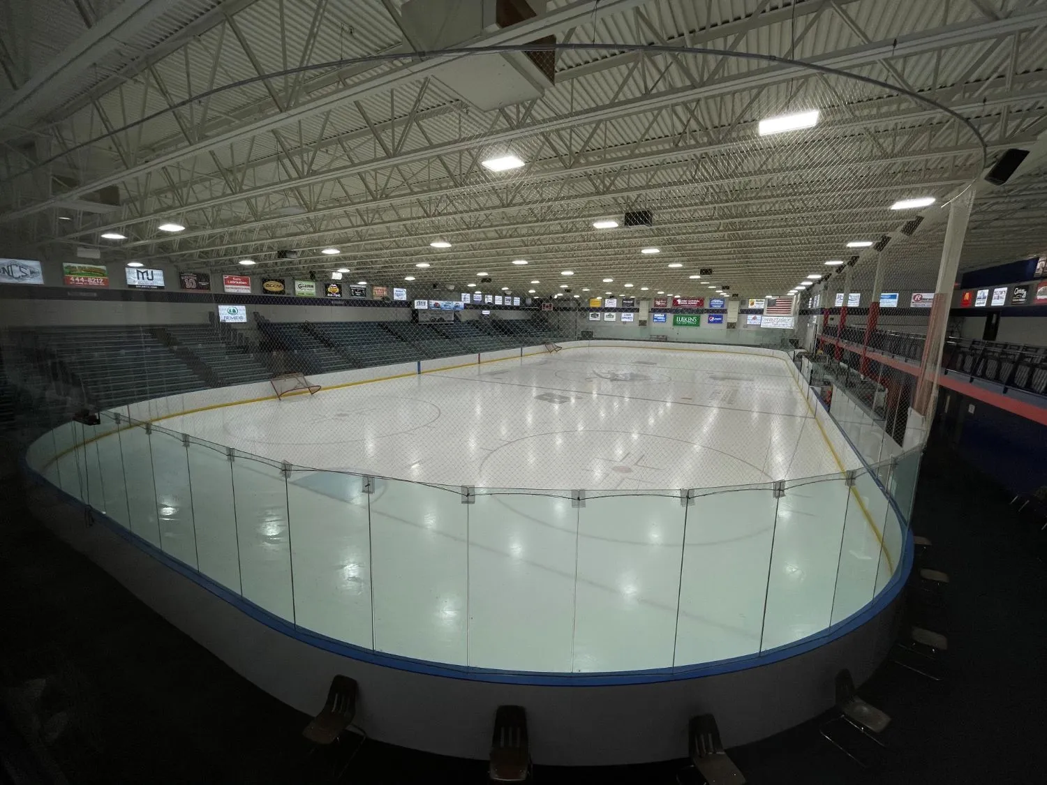 A wide view of an empty, freshly surfaced sheet behind the glass, with sponsor boards along the walls under the steel roof trusses. — Bemidji Community Arena