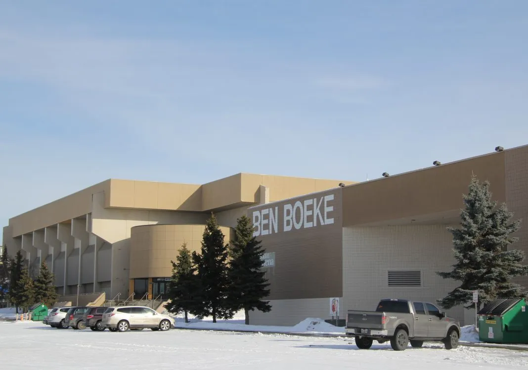 Front corner of the tan Ben Boeke Ice Arena building with large BEN BOEKE wall lettering, parking lot with cars and a pickup truck in snow, evergreen trees along the entrance. — Ben Boeke Ice Arena