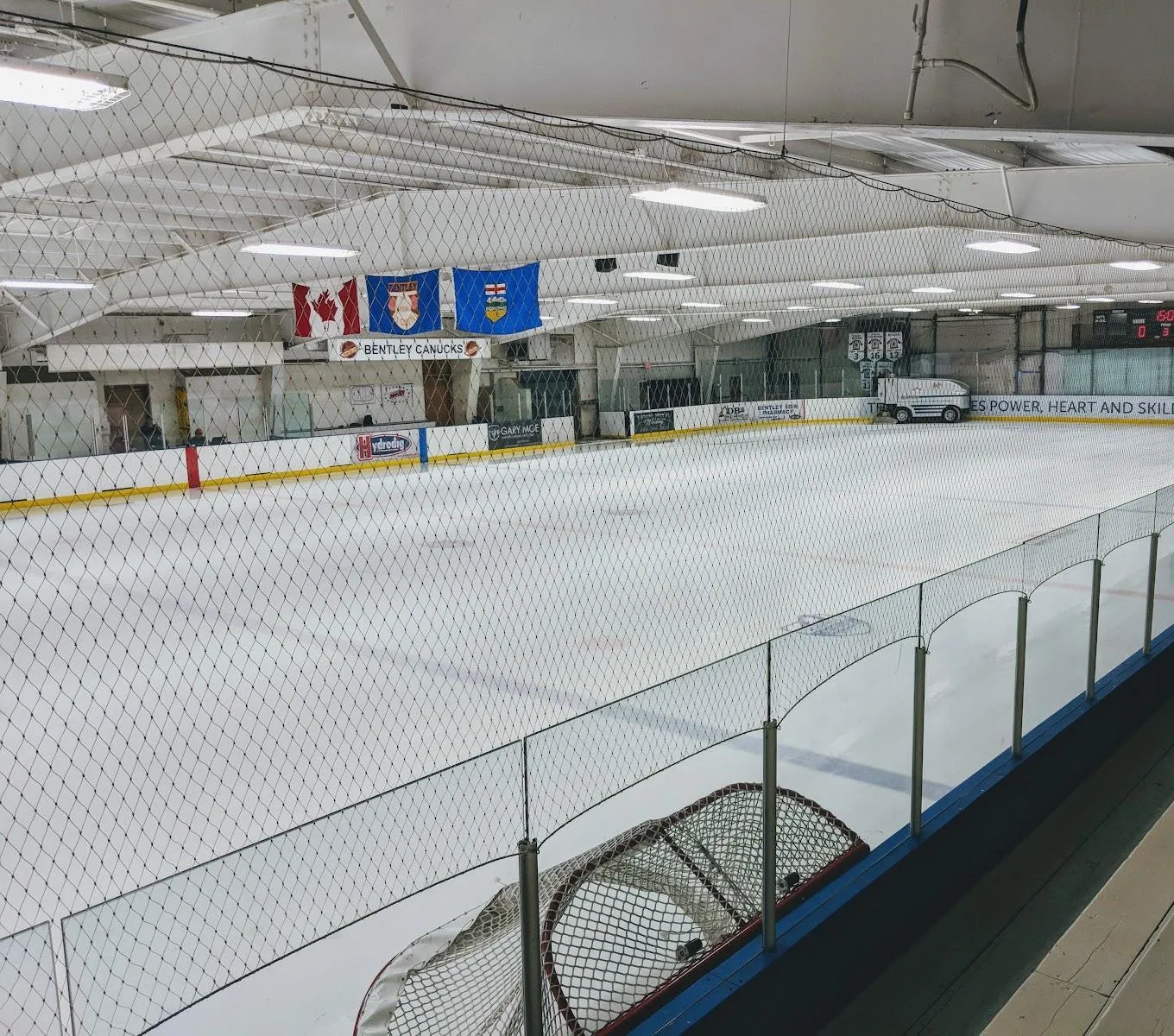 El hielo vacío de Bentley Arena desde las gradas, bajo las banderas de Canadá, Bentley y Alberta y una manta de los Bentley Canucks. — Bentley Arena