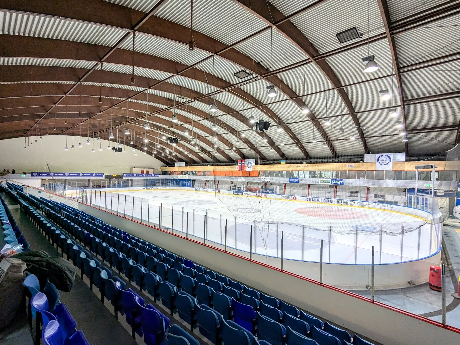 The empty ice sheet in Bergenshallen seen from the stand, under a curved timber roof, with blue seats along the near side. — Bergenshallen