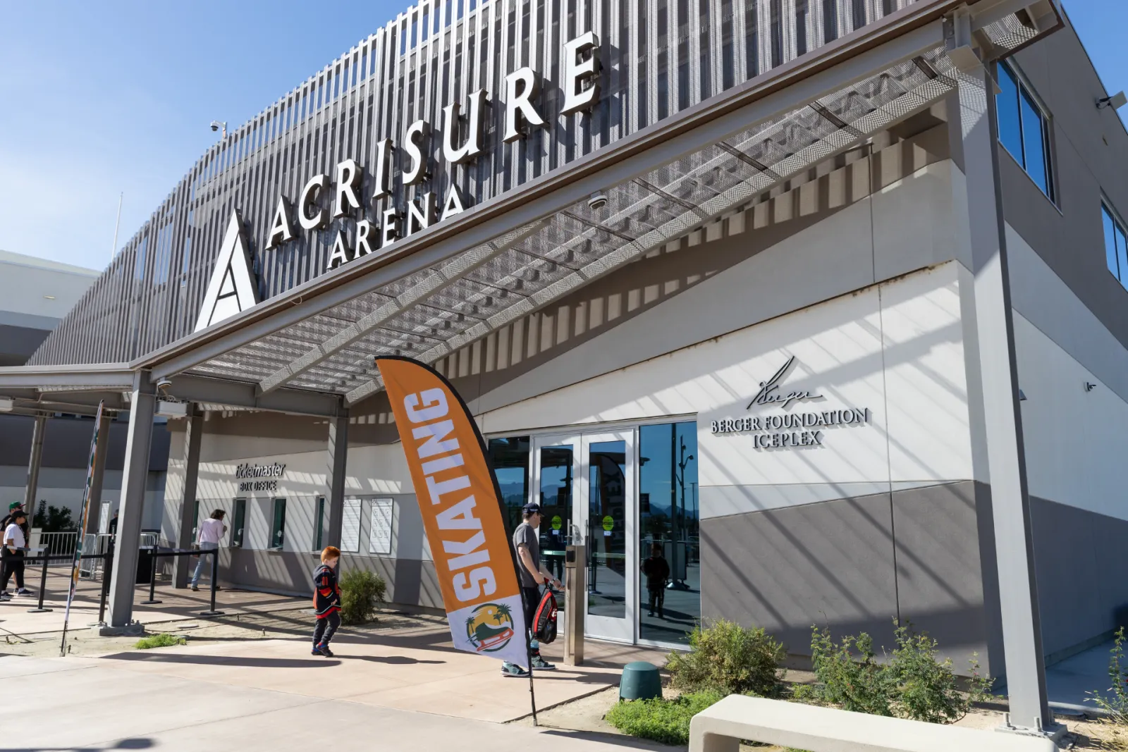 Entrance facade shared with Acrisure Arena, with a separate 'Berger Foundation Iceplex' sign and glass doors under an orange SKATING banner, a few people walking in. — Berger Foundation Iceplex