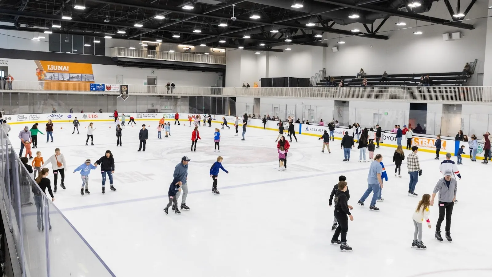A crowded public session on a wide sheet, skaters spread across the ice beneath a dark steel roof with sponsor boards along the far wall. — Berger Foundation Iceplex