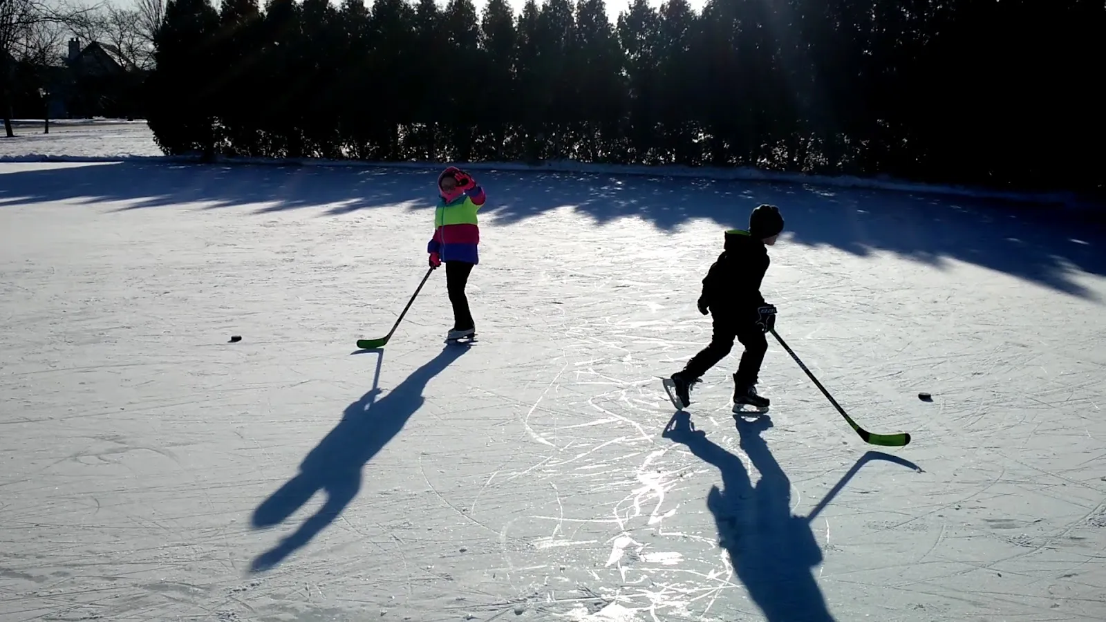 Two skaters on the backyard-style park rink in low winter sun, seen from a distance — Betty Russell Community Park Ice Rink
