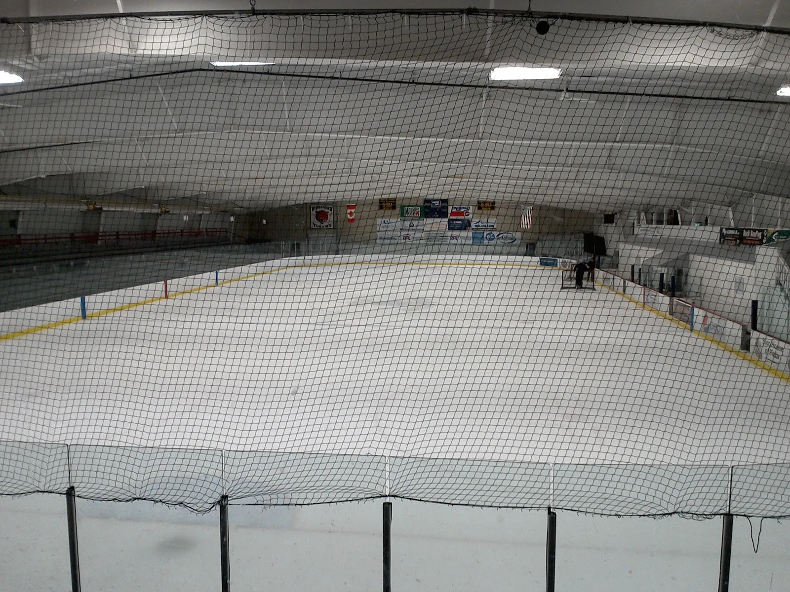 The empty ice sheet at Biddeford Ice Arena seen through the end netting, with the boards, flag and roof trusses — Biddeford Ice Arena