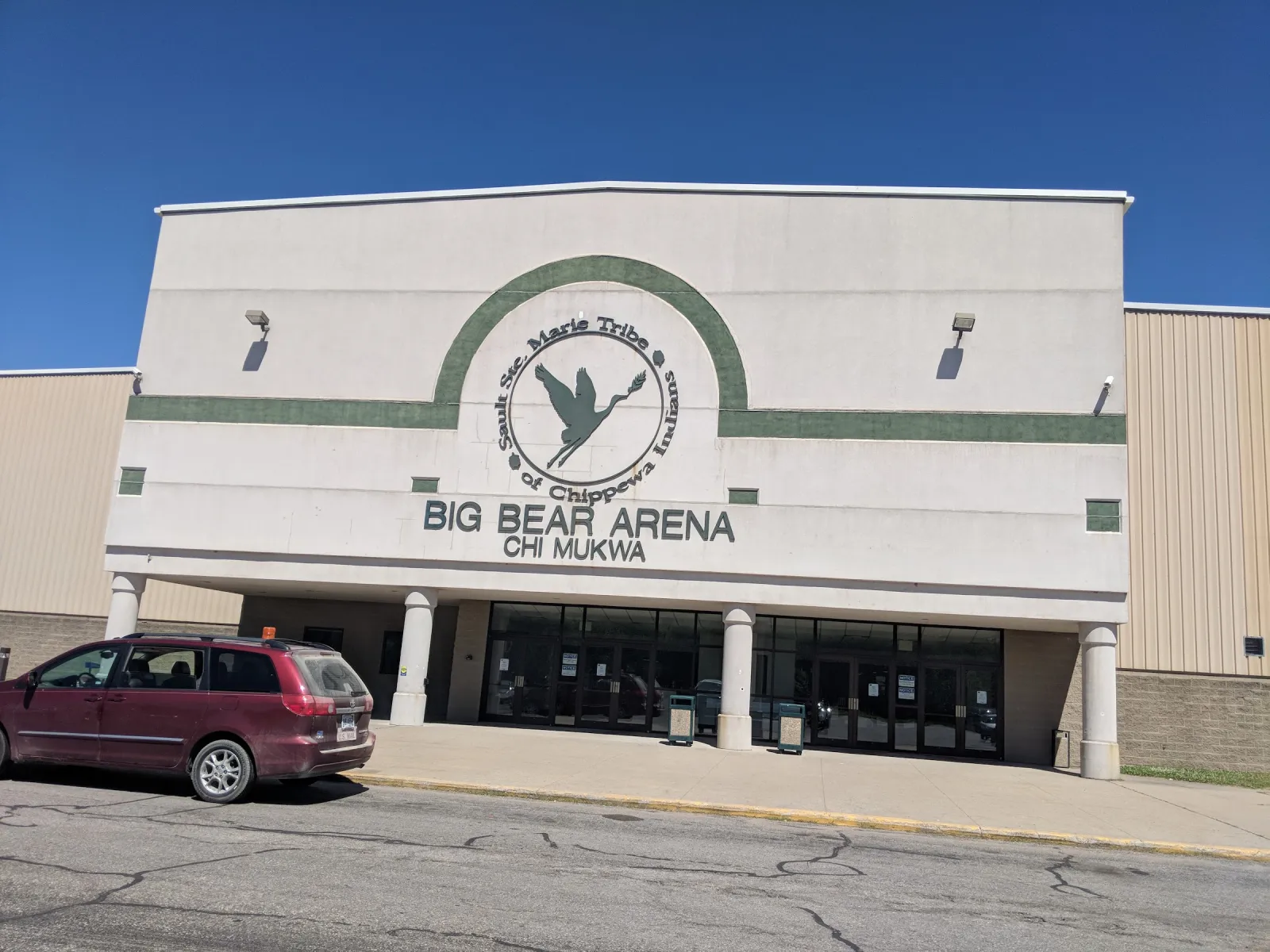 La fachada blanca de Big Bear Arena, con el escudo de la Sault Tribe y los nombres Big Bear Arena y Chi Mukwa sobre la entrada de cristal. — Big Bear Arena