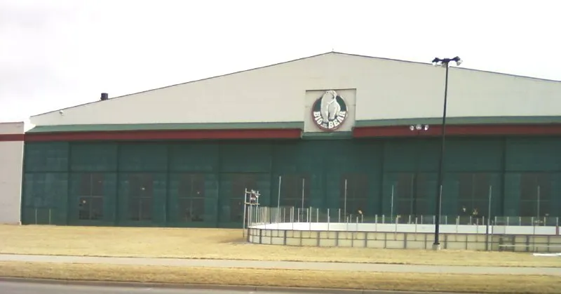 Front facade of Big Bear Ice Arena, a former Lowry Air Force Base hangar, with the round Big Bear logo sign on the gable end and a fenced outdoor area in front. — Big Bear Ice Arena