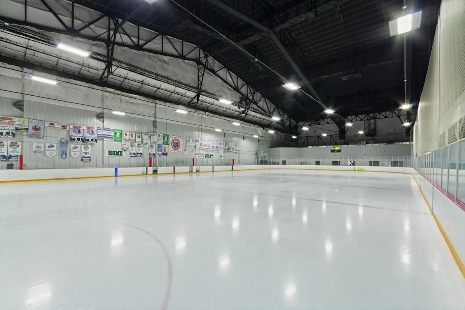 An empty Big Bear sheet under black steel trusses, with tournament banners on the long wall and the scoreboard at the far end — Big Bear Ice Arena