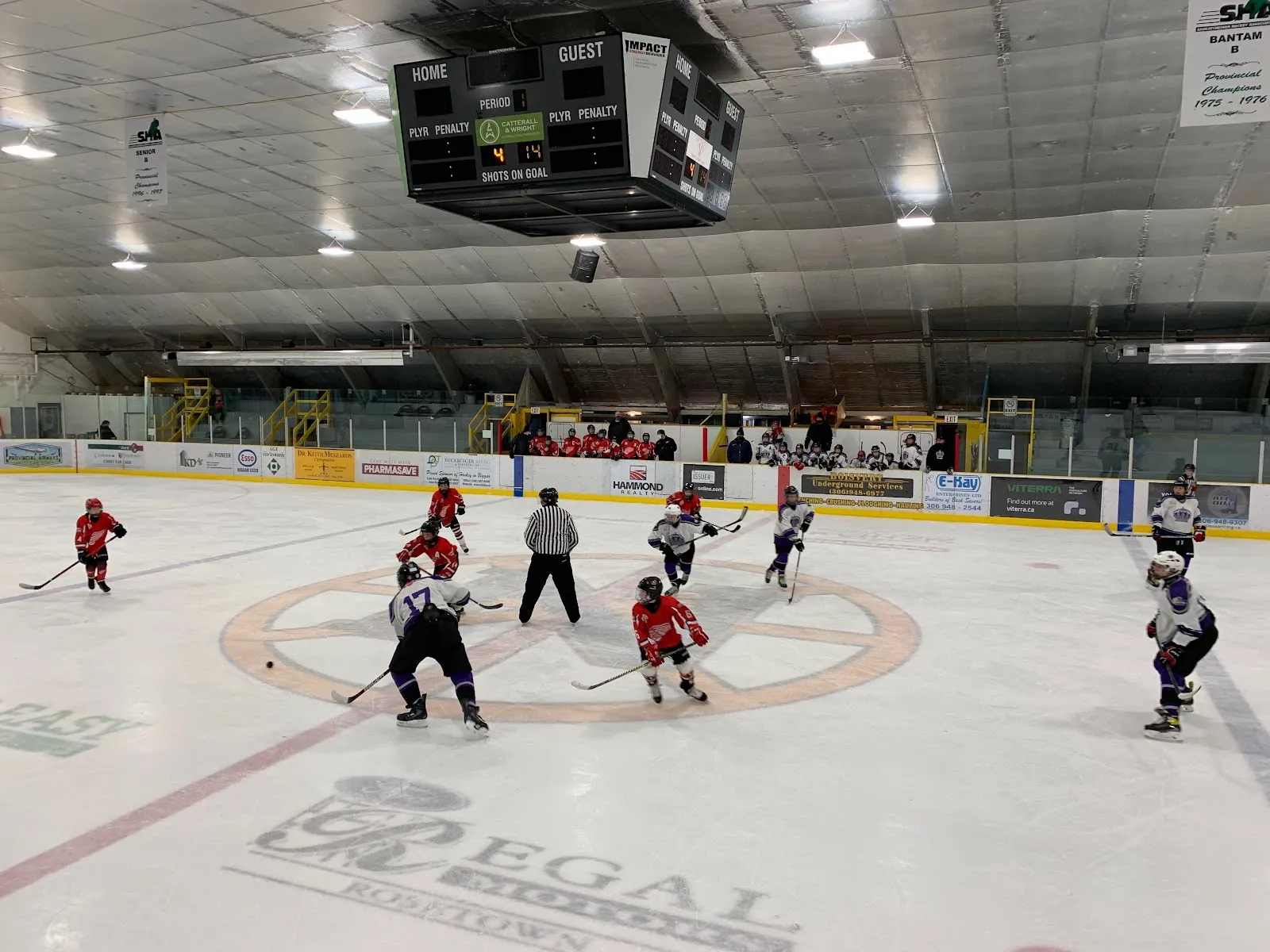 A youth game at the Jubilee Stadium seen from behind centre: the arched roof, scoreboard over centre ice, sponsor boards and spectators along the far side. — Biggar Jubilee Stadium