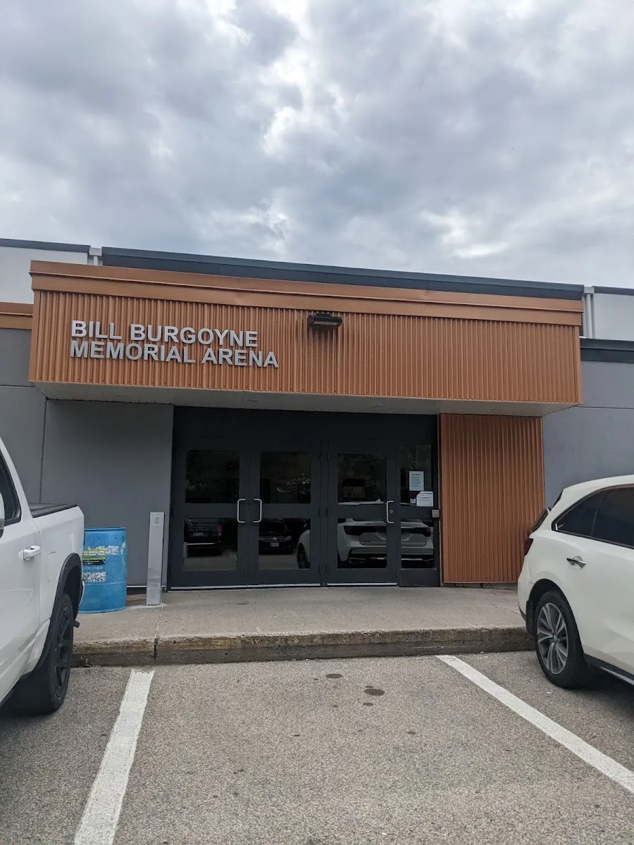La entrada de la arena con la leyenda BILL BURGOYNE MEMORIAL ARENA en letras en relieve sobre una marquesina con paneles de madera encima de las puertas de vidrio, y autos estacionados al frente. — Bill Burgoyne Memorial Arena